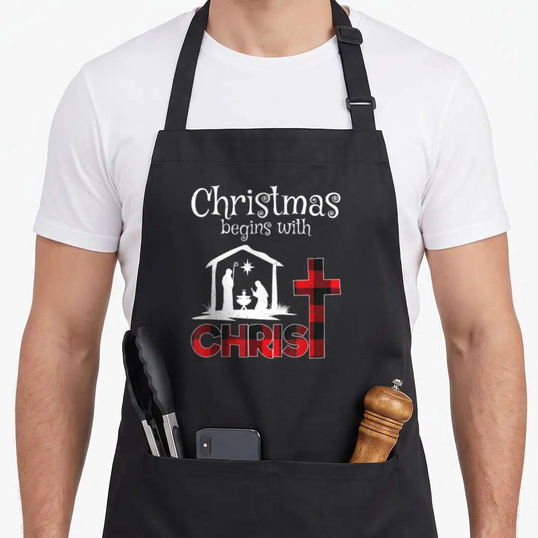 Begin buffalo plaid christmas apron - Nativity scene and cross with red black plaid text reading Christmas begins — shown on a black cotton canvas apron against a white background