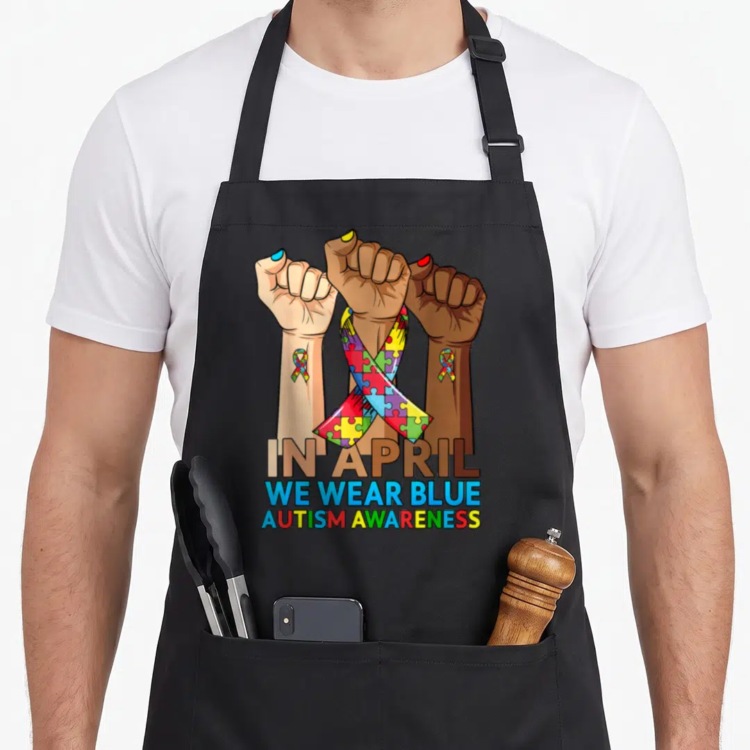 Hand april apron - Three raised fists with puzzle ribbons and text IN APRIL WE WEAR BLUE — shown on a black cotton canvas apron against a white background
