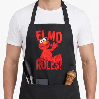 Rule elmo apron - Red cartoon character between ELMO and RULES text in orange and white — shown on a black cotton canvas apron against a white background