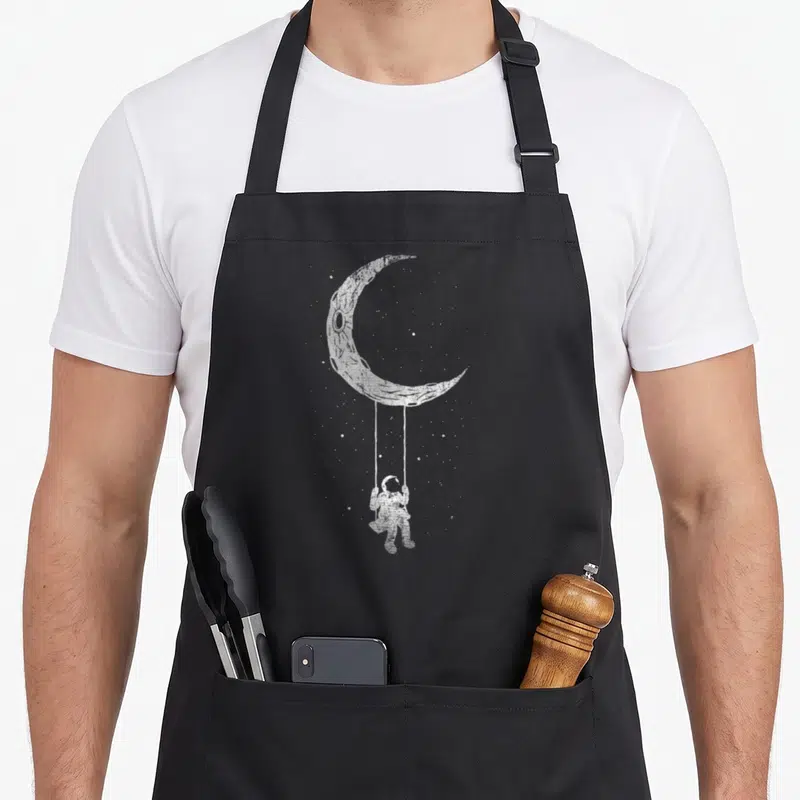 Space funny moon astronomy lovers apron - White line art of astronaut on swing hanging from crescent moon surrounded — shown on a black cotton canvas apron against a white background