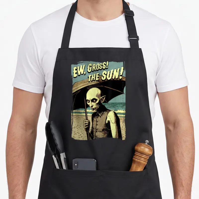 Gross goth apron - Vampire holding umbrella on beach with text EW GROSS THE SUN in distressed yellow and black — shown on a black cotton canvas apron against a white background