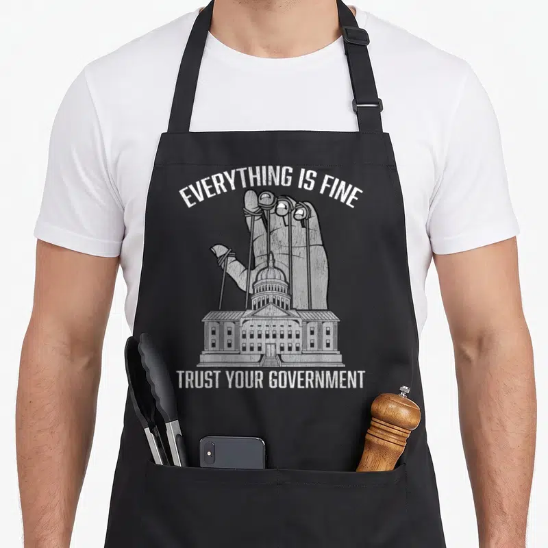 Fine conspiracy apron - Giant hand puppeting government building with Everything Is Fine Trust Your Government text — shown on a black cotton canvas apron against a white background