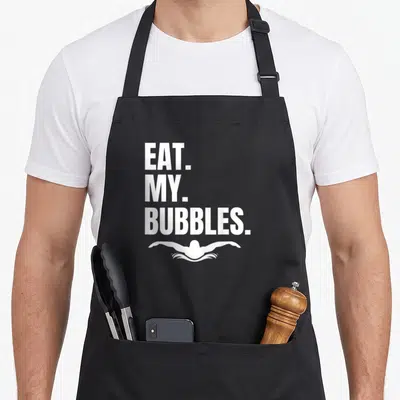 Eat swimmer apron - EAT MY BUBBLES text above white swimmer silhouette in minimalist style — shown on a black cotton canvas apron against a white background
