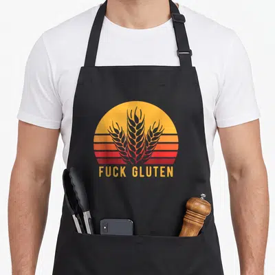 Grain celiac awareness apron - Retro sunset graphic with three grey wheat stalks and yellow FUCK GLUTEN text below — shown on a black cotton canvas apron against a white background