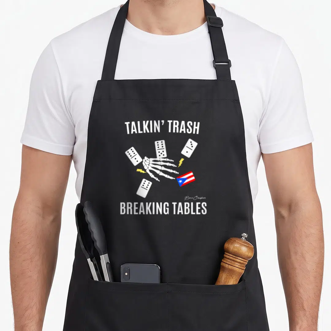 Puerto dominoes apron - Skeleton hand holding dominoes with talkin trash breaking tables text and flag — shown on a black cotton canvas apron against a white background