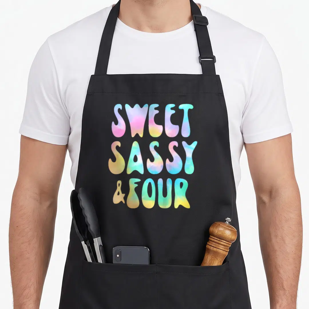 Four sassy apron - Colorful bubble letters spelling SWEET SASSY & FOUR in tie dye gradient — shown on a black cotton canvas apron against a white background