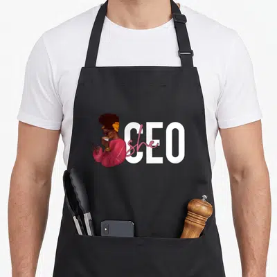 She small business aprons - Cartoon woman in pink sweater holding coffee next to white CEO text with pink she script — shown on a black cotton canvas apron against a white background