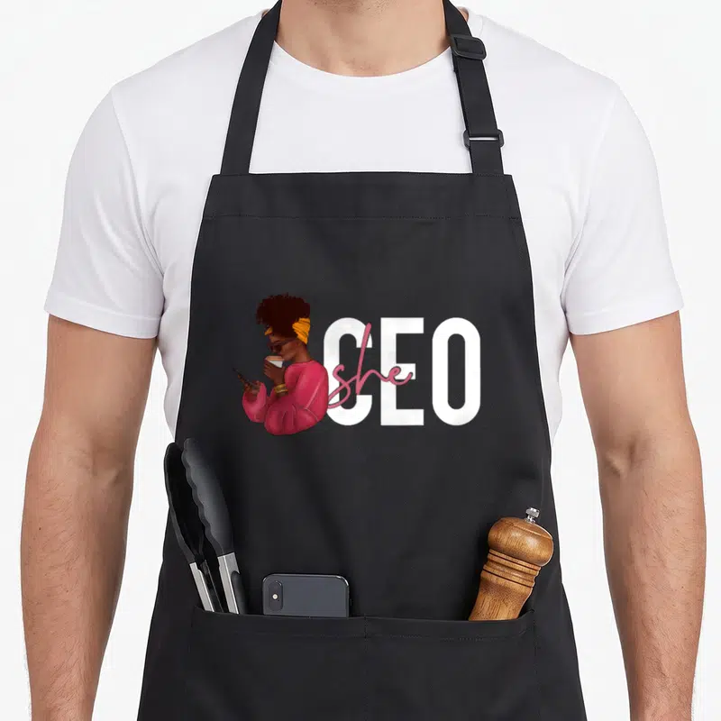 She small business aprons - Cartoon woman in pink sweater holding coffee next to white CEO text with pink she script — shown on a black cotton canvas apron against a white background