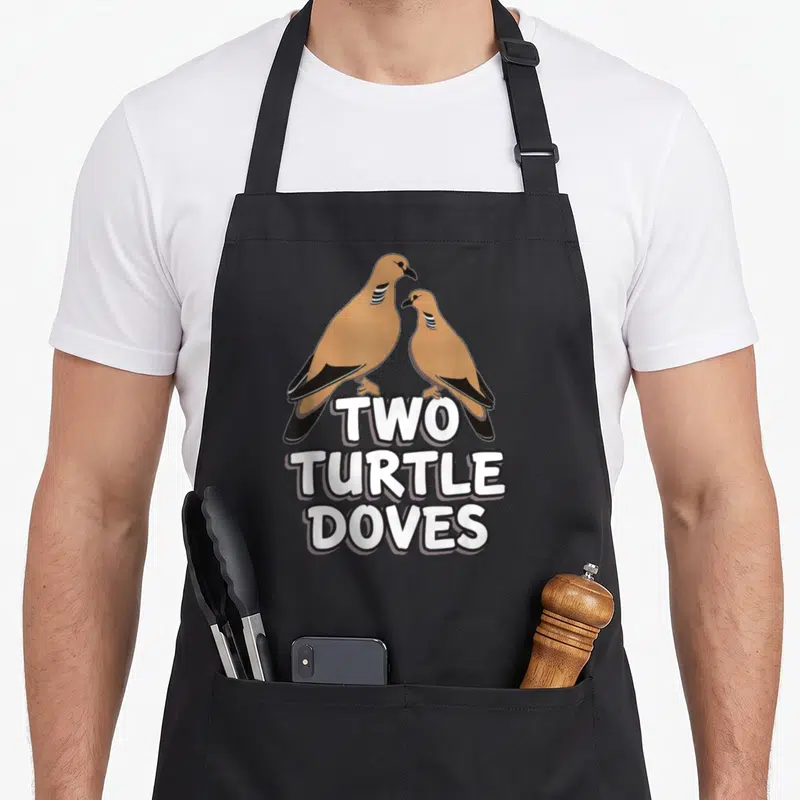 Song christmas apron - Two tan cartoon birds standing above stacked white text reading TWO TURTLE DOVES — shown on a black cotton canvas apron against a white background