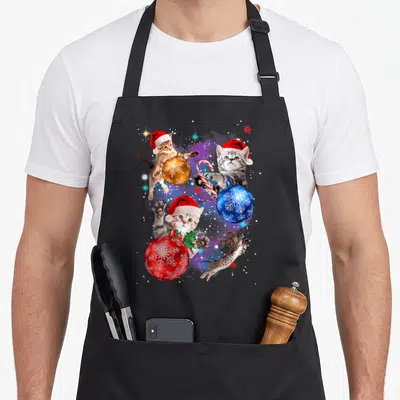Space christmas cat apron - Four kittens with Santa hats and antlers playing among red blue and gold ornaments — shown on a black cotton canvas apron against a white background