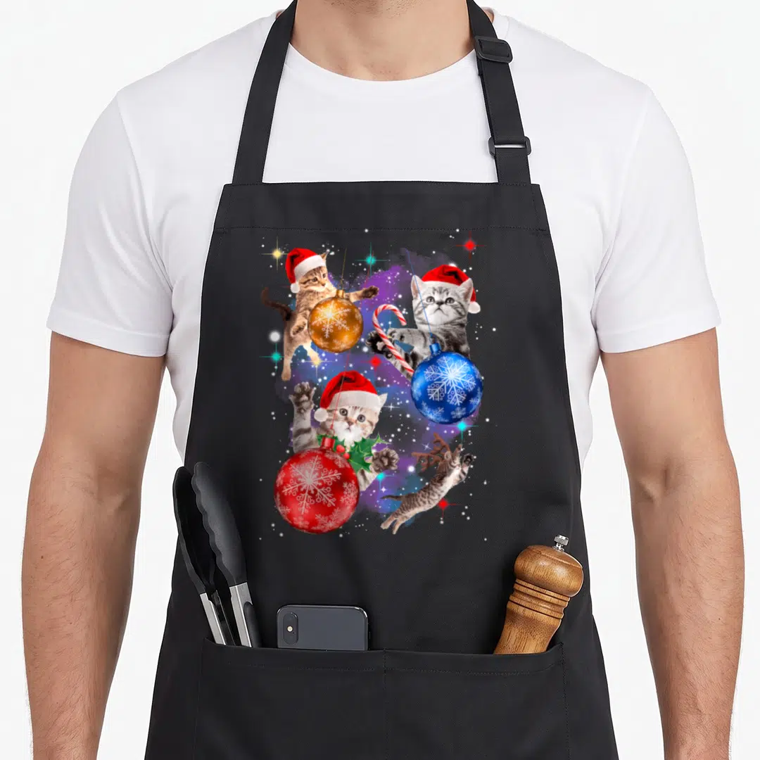 Space christmas cat apron - Four kittens with Santa hats and antlers playing among red blue and gold ornaments — shown on a black cotton canvas apron against a white background