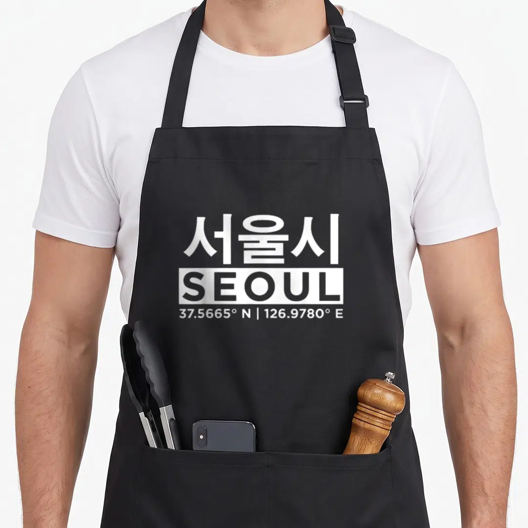 Funny seoul south korea apron - White stacked text showing Seoul in Korean and English with coordinates 37.5665 N — shown on a black cotton canvas apron against a white background