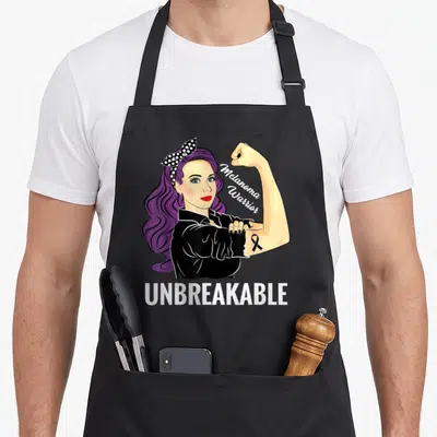 Product melanoma awareness apron - Cartoon woman with purple hair flexing arm showing black ribbon and UNBREAKABLE text — shown on a black cotton canvas apron against a white background