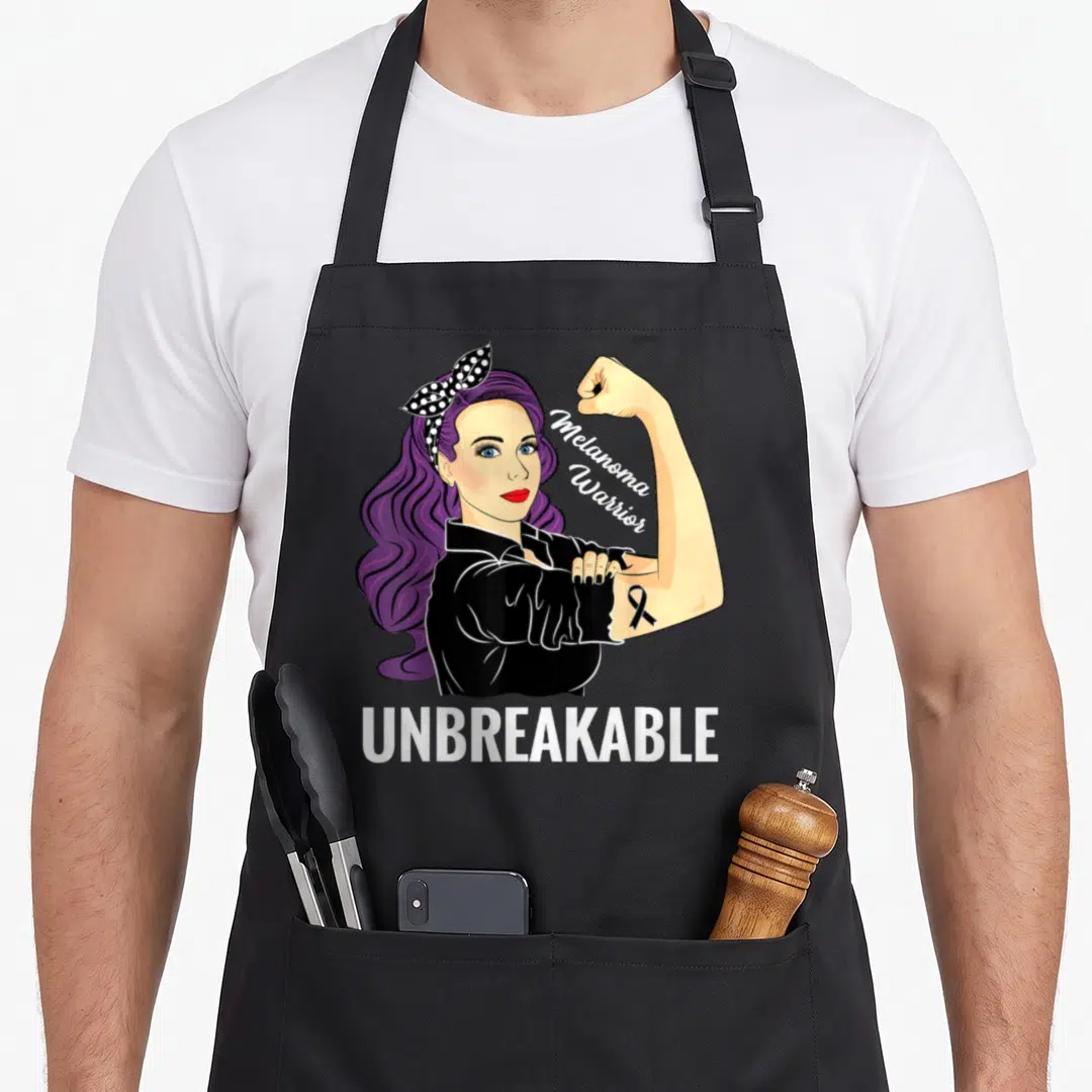 Product melanoma awareness apron - Cartoon woman with purple hair flexing arm showing black ribbon and UNBREAKABLE text — shown on a black cotton canvas apron against a white background