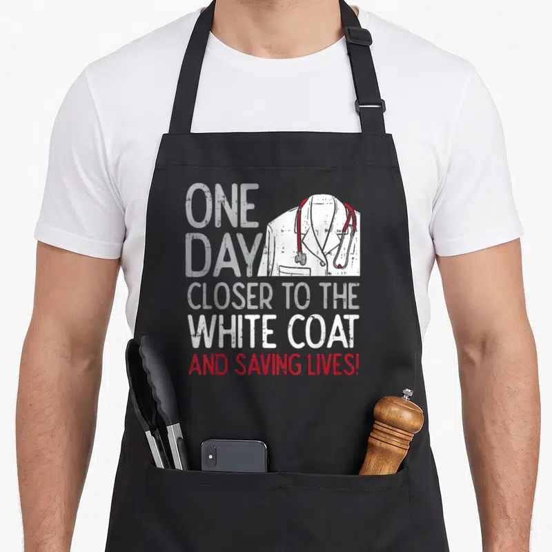 Future lifesaver doctor apron - Distressed text ONE DAY CLOSER TO THE WHITE COAT next to a white coat with red — shown on a black cotton canvas apron against a white background