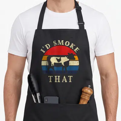 Smoke smoker meat aprons - Retro cow silhouette against striped sun with I'D SMOKE, THAT text — shown on a black cotton canvas apron against a white background