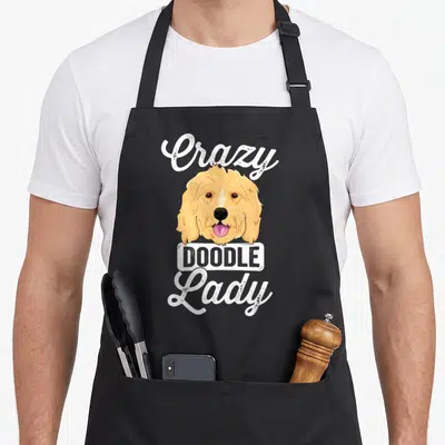 Doodle dog cooking apron - Cartoon tan dog head with pink tongue and Crazy Doodle Lady text — shown on a black cotton canvas apron against a white background