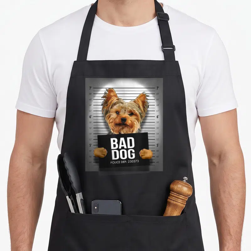 Mugshot yorkie apron - Dog holding black sign with Bad Dog Police Dep text in front of height chart — shown on a black cotton canvas apron against a white background