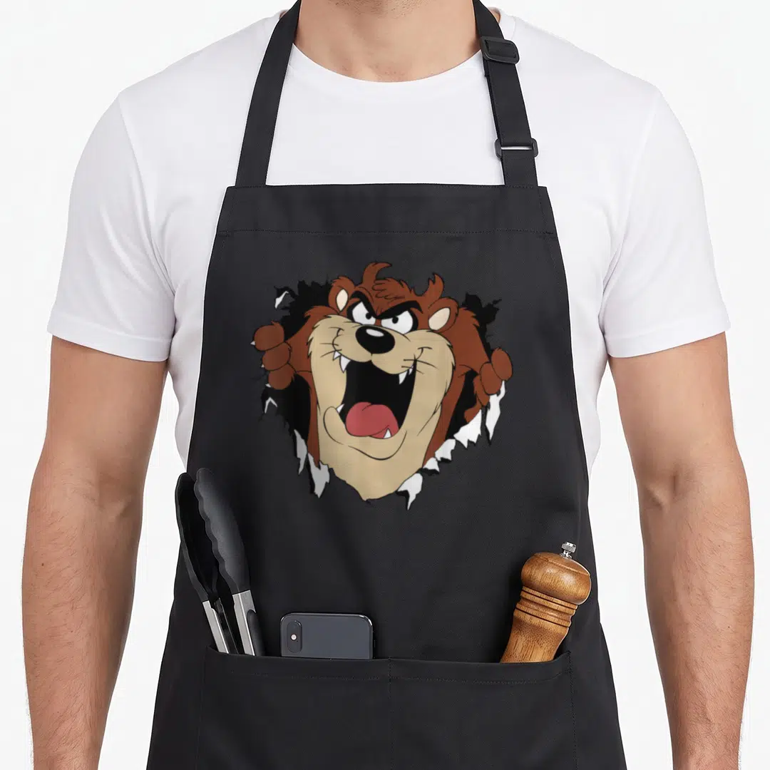 Taz looney tune apron - Cartoon bear bursting through torn black and white fabric with brown fur and tan muzzle — shown on a black cotton canvas apron against a white background