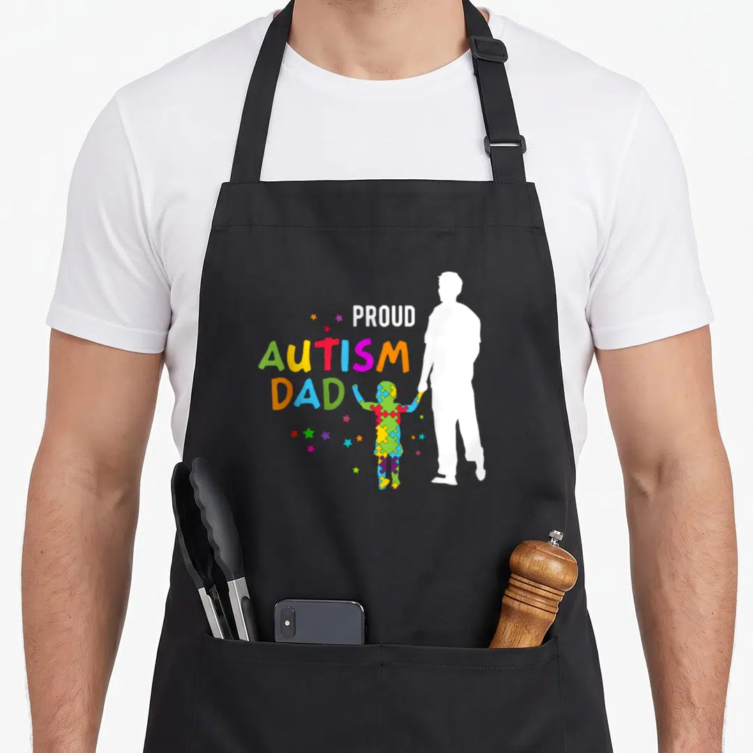 Awareness proud autism dad apron - Stacked PROUD AUTISM DAD text next to white father silhouette holding colorful — shown on a black cotton canvas apron against a white background