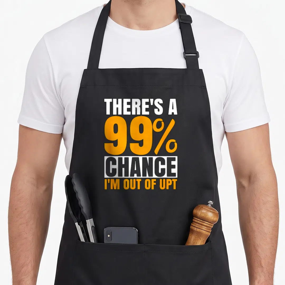 Chance swagazon apron - Stacked text reading THERE'S A 99% CHANCE I'M OUT OF UPT in white orange and grey — shown on a black cotton canvas apron against a white background