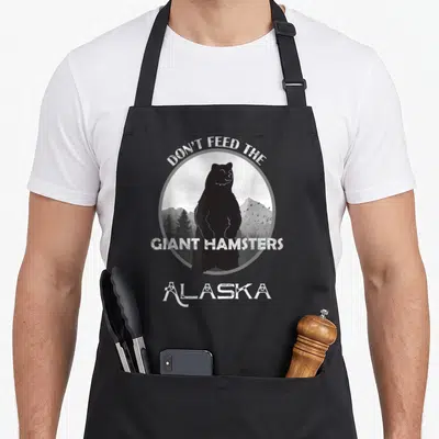 Alaskan grizzly bear apron - Black bear silhouette in grey circle with Don't Feed The Giant Hamsters Alaska text — shown on a black cotton canvas apron against a white background