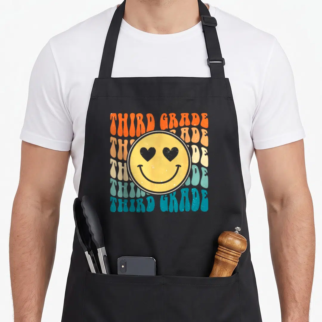 Boho teacher apron - Yellow smiley face with heart eyes over six stacked THIRD GRADE text lines in orange blue beige — shown on a black cotton canvas apron against a white background