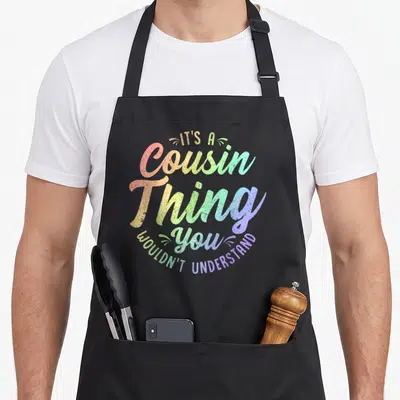 Cousin family reunion aprons - Rainbow text reading It's a Cousin Thing You Wouldn't Understand in stacked script — shown on a black cotton canvas apron against a white background