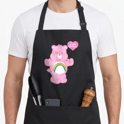 Funny cheer apron - Pink bear with rainbow belly and Care Bears heart logo in cartoon style — shown on a black cotton canvas apron against a white background