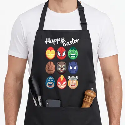 Funny easter egg apron - Nine colorful character eggs in 3x3 grid under arched Happy Easter text in cartoon style — shown on a black cotton canvas apron against a white background