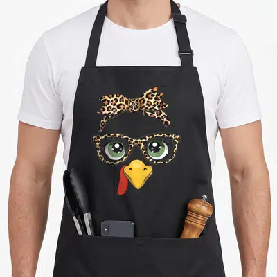Thanksgiving leopard print apron - Cartoon turkey face with leopard print glasses, green eyes, yellow beak, and red — shown on a black cotton canvas apron against a white background