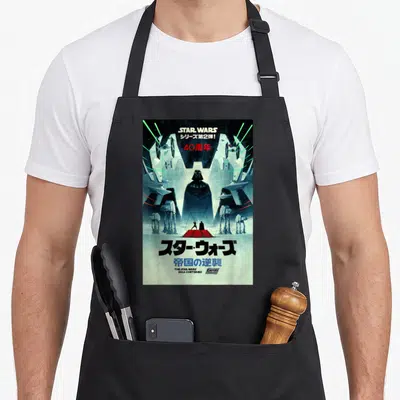 Empire 40th apron - Retro Star Wars poster with Darth Vader, AT-ATs, TIE fighters, and Japanese anniversary text — shown on a black cotton canvas apron against a white background
