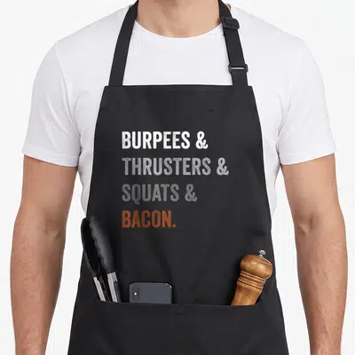 Burpee bacon apron - Stacked text reading Burpees Thrusters Squats Bacon in white grey and orange — shown on a black cotton canvas apron against a white background