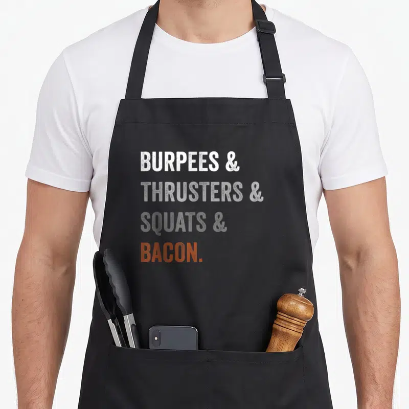 Burpee bacon apron - Stacked text reading Burpees Thrusters Squats Bacon in white grey and orange — shown on a black cotton canvas apron against a white background