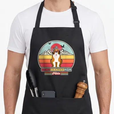 Retro saint bernard apron - Saint Bernard dog with glasses and bandana in retro circle with mountains and text banners — shown on a black cotton canvas apron against a white background