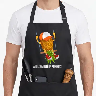 Gear pineapple apron - Cartoon pineapple in cap and red shoes sitting on swing with Will Swing If Pushed text below — shown on a black cotton canvas apron against a white background