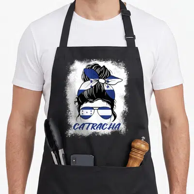 Catracha honduran apron - Stylized woman head with bun and sunglasses in blue white flag pattern with CATRACHA text — shown on a black cotton canvas apron against a white background