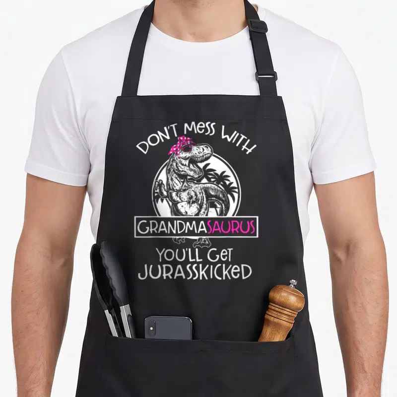 Mess grandma apron - Dinosaur with pink glasses and bandana, text arching over and stacking below in white and pink — shown on a black cotton canvas apron against a white background