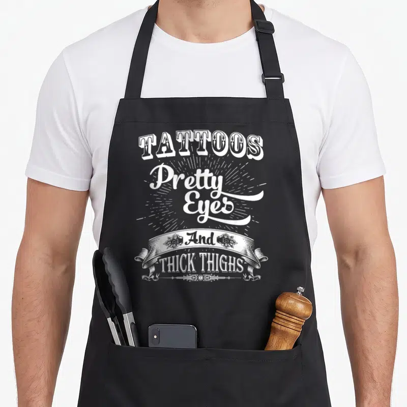 Eye tattoo apron - White stacked text Tattoos Pretty Eyes And THICK THIGHS with sunburst and ribbon — shown on a black cotton canvas apron against a white background