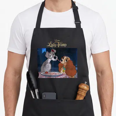 Iconic lady and the tramp apron - Disney Lady and the Tramp text above two cartoon dogs sharing spaghetti — shown on a black cotton canvas apron against a white background