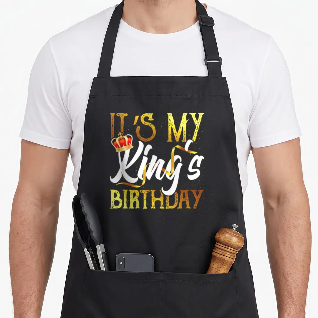 Proud kings apron - Gold and white text reading It's My King's Birthday with red and gold crown — shown on a black cotton canvas apron against a white background