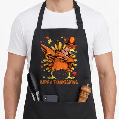 Cute dabbing thanksgiving day aprons - Cartoon turkey with pilgrim hat and heart sunglasses surrounded by fall leaves — shown on a black cotton canvas apron against a white background