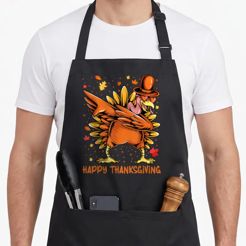 Cute dabbing thanksgiving day aprons - Cartoon turkey with pilgrim hat and heart sunglasses surrounded by fall leaves — shown on a black cotton canvas apron against a white background