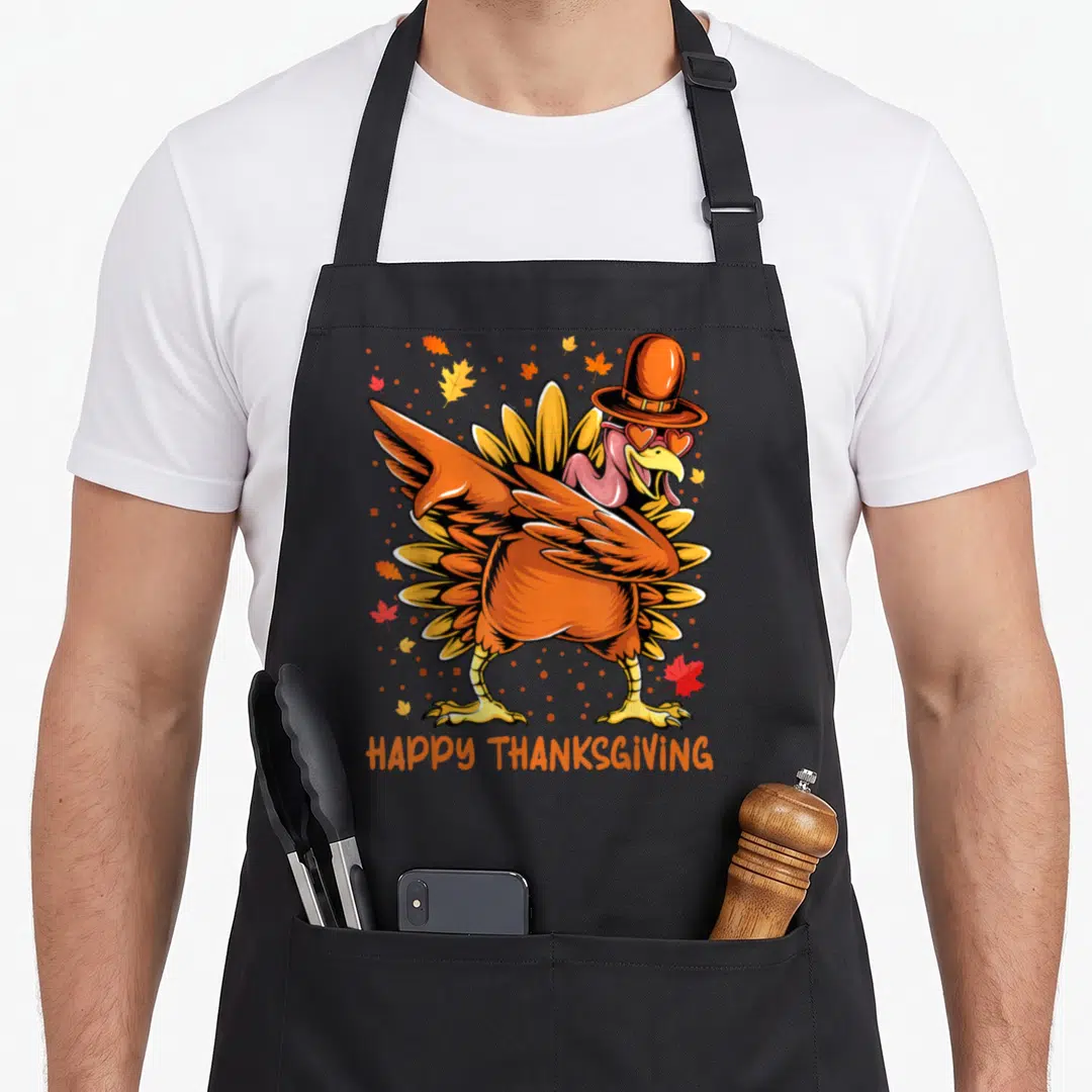Cute dabbing thanksgiving day aprons - Cartoon turkey with pilgrim hat and heart sunglasses surrounded by fall leaves — shown on a black cotton canvas apron against a white background