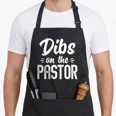 Dib wife apron - White stacked text Dibs on the PASTOR in script and block fonts with distressed finish — shown on a black cotton canvas apron against a white background