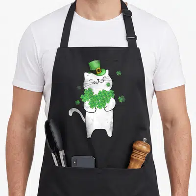 Cat st patrick day aprons - White cartoon cat wearing green hat and holding cluster of shamrocks — shown on a black cotton canvas apron against a white background