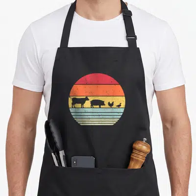 Chicken farm aprons - Retro circular sunset with cow, pig, chicken, and duck silhouettes on red orange yellow blue — shown on a black cotton canvas apron against a white background