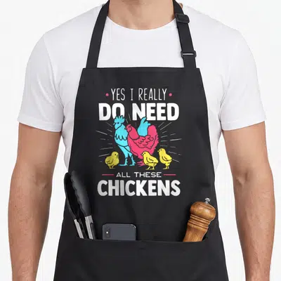 Chicken do i need an apron - Cartoon blue rooster pink hen and three yellow chicks with white text reading yes i really — shown on a black cotton canvas apron against a white background