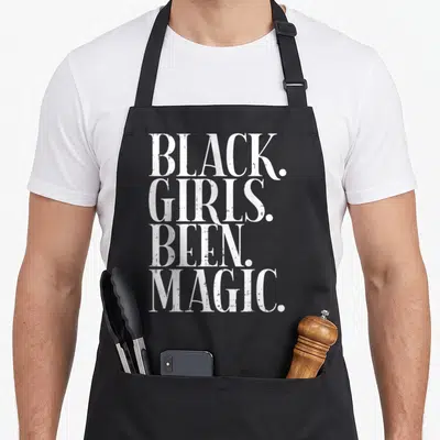 Magic african american aprons - White distressed text reading BLACK. GIRLS. BEEN. MAGIC. stacked vertically on black — shown on a black cotton canvas apron against a white background