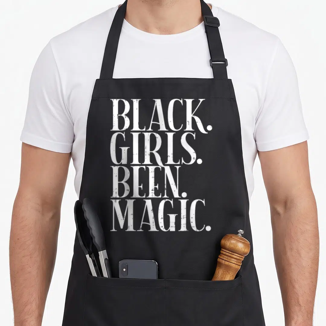 Magic african american aprons - White distressed text reading BLACK. GIRLS. BEEN. MAGIC. stacked vertically on black — shown on a black cotton canvas apron against a white background